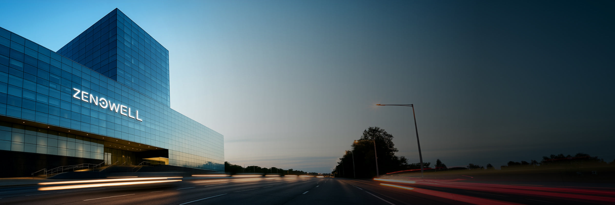 ZenoWell office building with illuminated logo, viewed from the road as vehicles pass by during sunset.
